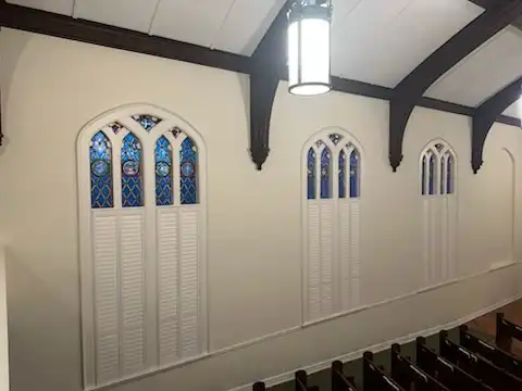 Arched stained glass church windows with custom white shutters, seen from the sanctuary balcony