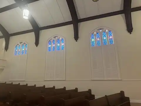 Three arched stained glass windows in a church sanctuary fitted with custom white shutters below the glass