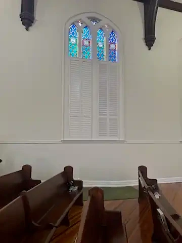 Tall arched stained glass church window with custom white plantation shutters on the lower section
