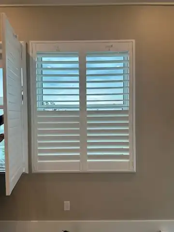 White plantation shutter panel swung open beside a closed panel, with a water view through the louvers