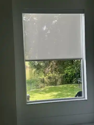 Light filtering roller shade raised halfway on a window overlooking a green backyard and fence