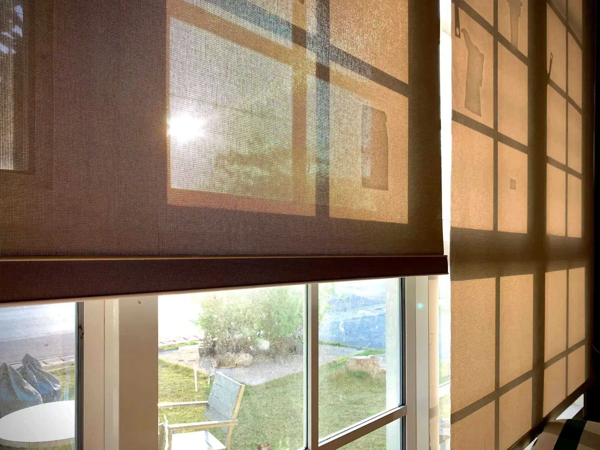 Roller shade half lowered over a sunlit window facing a garden