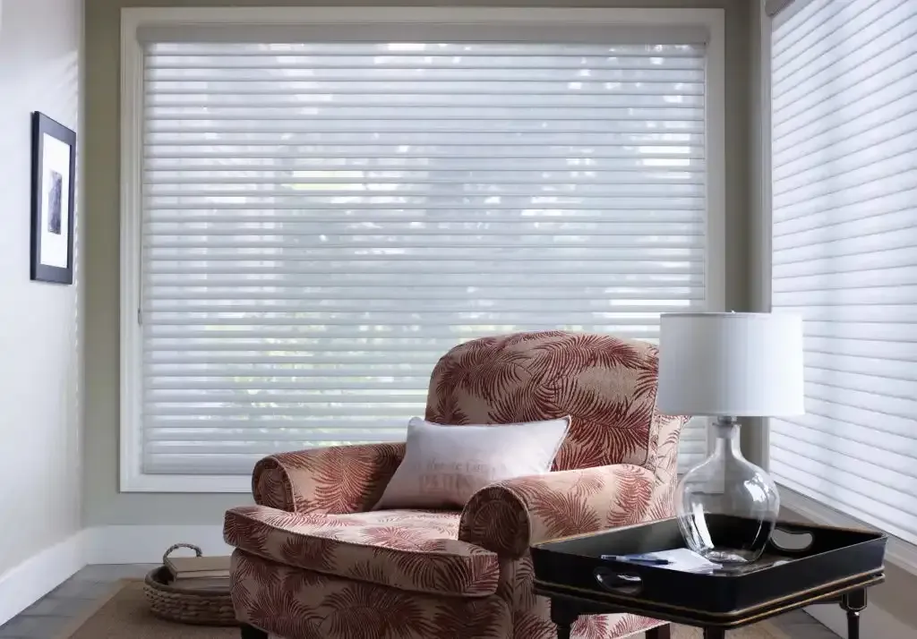 Armchair in front of a window with sheer blinds. A table with lamp and decor sits beside the chair.