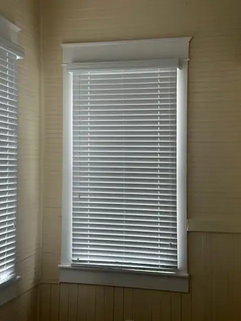 White horizontal blinds lowered on a tall cottage window with painted wood trim and beadboard walls