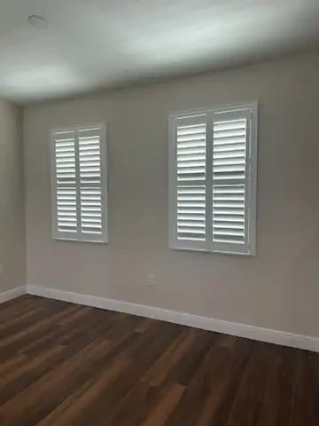 Two bedroom windows fitted with matching white plantation shutters above dark wood flooring
